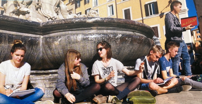 Fünf Jugendliche sitzen im Schneidersitz vor einem Brunnen, ein Jugendlicher steht. Vier von ihnen haben ein Skizzenbuch in der Hand oder im Schoß. 